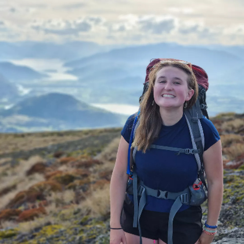 The image shows a young woman smiling at the camera while hiking. She is wearing a backpack and appears to be on a mountain, with a scenic view of mountains and a lake in the background. The lighting suggests it's daytime, and the overall mood is positive and adventurous.
